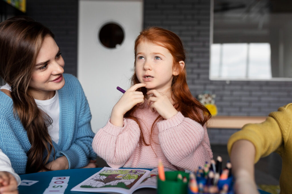 Young girl practicing speech sounds by touching her chin during a therapy activity, with a speech-language pathologist sitting nearby.